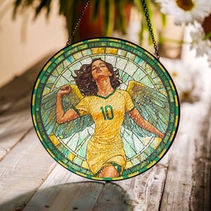 以下が含まれることがあります： チェーンで吊るされたステンドグラスの窓。窓には、10番の黄色いジャージを着た翼のある女性が描かれています。背景は緑、青、黄色のモザイクです。