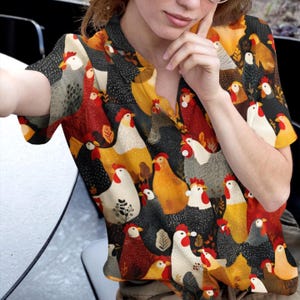 Puede incluir: Una camisa abotonada con un patrón repetido de gallinas coloridas en varias poses. Las gallinas son en tonos de rojo, amarillo, blanco y negro sobre un fondo oscuro. La camisa tiene mangas cortas y cuello.