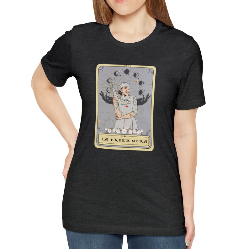 May include: A black t-shirt featuring a tarot card design with a woman in a nurse's uniform holding a syringe. The card is titled "La Enfermera" in Spanish.