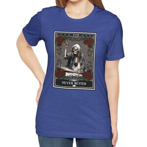 Pu&ograve; includere: Una t-shirt blu reale con un design di carta dei tarocchi che presenta uno scheletro con un viso di teschio, seduto con una tazza in mano. La carta &egrave; etichettata come "XVII" e "NEVER BETTER".