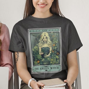 May include: Dark gray t-shirt featuring a tarot card design with the text "THE GREEN WITCH." The illustration depicts a woman with long blonde hair surrounded by plants and flowers. The card is framed with a green border and the Roman numeral XVII.