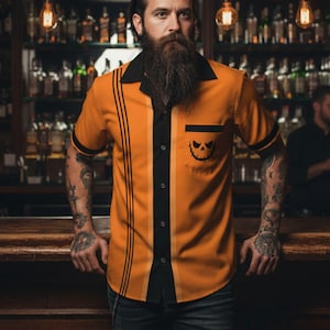 Op de afbeelding: Oranje overhemd met korte mouwen met zwarte details en een Halloween-thema. Het shirt heeft een zwarte kraag, zak en verticale strepen. Een pompoengezicht is op de zak gedrukt. Gemaakt van lichtgewicht materiaal.