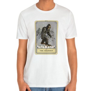 Pode incluir: Camiseta branca com um gr&aacute;fico de uma criatura Bigfoot em um estilo de carta de tar&ocirc; vintage. O cart&atilde;o &eacute; amarelo com uma borda dourada e apresenta o texto "THE BIGFOOT".