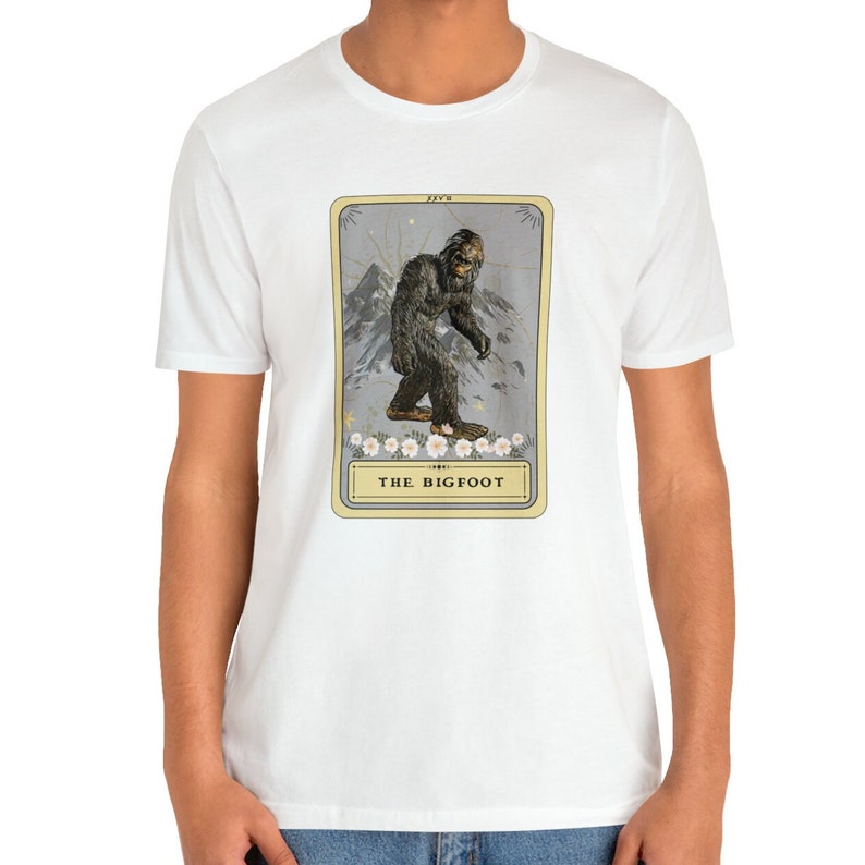 Pode incluir: Camiseta branca com um gr&aacute;fico de uma criatura Bigfoot em um estilo de carta de tar&ocirc; vintage. O cart&atilde;o &eacute; amarelo com uma borda dourada e apresenta o texto "THE BIGFOOT".