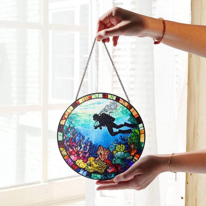 Op de afbeelding: Een rond glas-in-loodraam dat aan een ketting hangt. Het raam toont een duiker in silhouet die door een kleurrijk koraalrif zwemt.