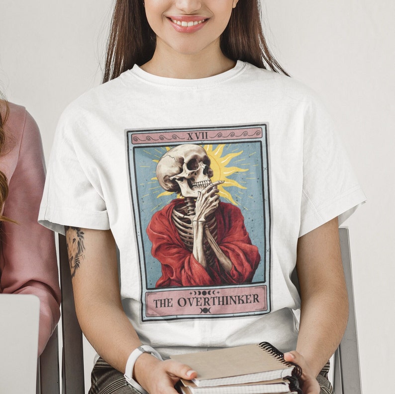May include: White t-shirt with a graphic of a skeleton in a red robe, holding a cigarette and looking thoughtful. The graphic is in the style of a tarot card, with the number XVIII and the text "THE OVERTHINKER" below the image.