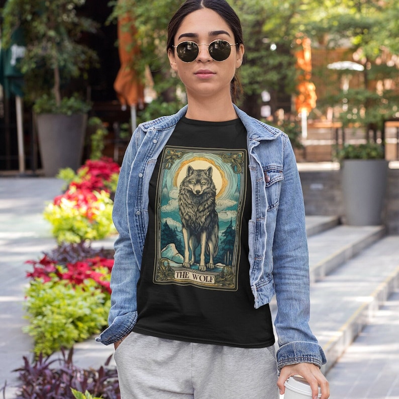 May include: A black t-shirt with a vintage-style illustration of a wolf howling at the moon. The illustration is surrounded by a decorative frame and the text "THE WOLF" is printed below the image.