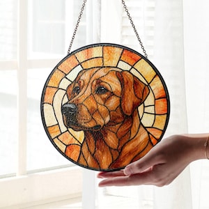 May include: A round stained glass window featuring a brown dog with a black nose and eyes. The dog is surrounded by a mosaic of orange, yellow, and brown glass pieces. The window is framed in black and hangs from a chain.