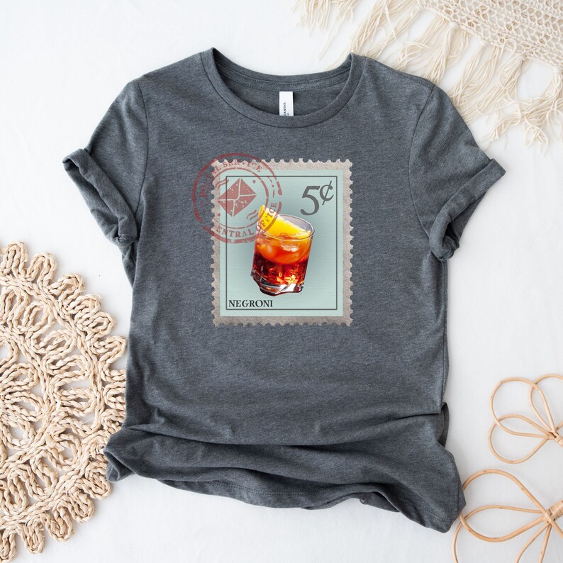 May include: A dark gray t-shirt with a vintage-style stamp design featuring a glass of Negroni with an orange slice. The stamp reads "5&cent;" and "NEGRONI".