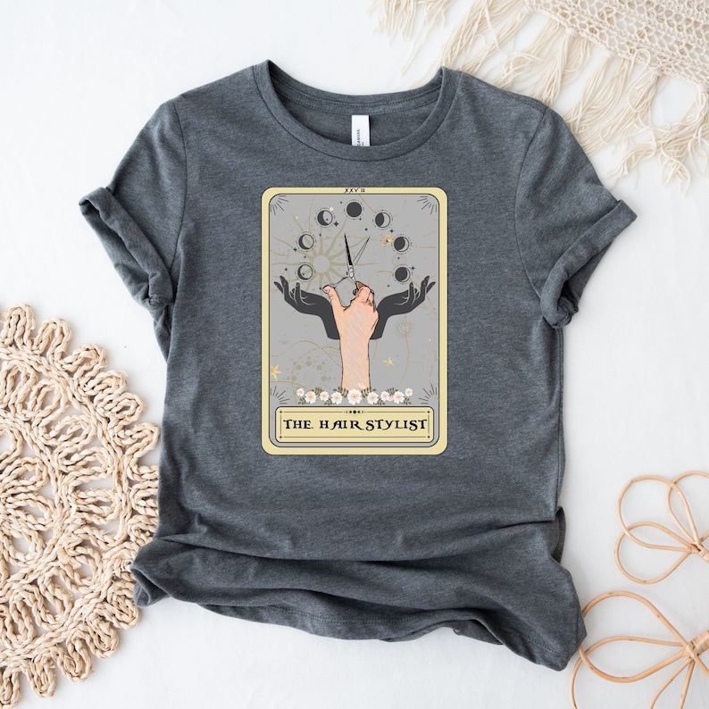 Puede incluir: Una camiseta gris con un gr&aacute;fico de un dise&ntilde;o de carta de tarot que presenta una mano sosteniendo tijeras y el texto "THE HAIR STYLIST".