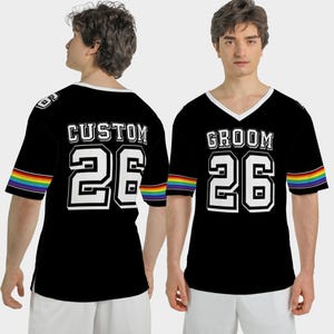 Puede incluir: Camiseta negra estilo jersey con ribete blanco y la palabra "GROOM" o "CUSTOM" en letras blancas. El n&uacute;mero "26" tambi&eacute;n es blanco. Las mangas tienen una franja arco&iacute;ris. La camiseta tiene cuello en V.