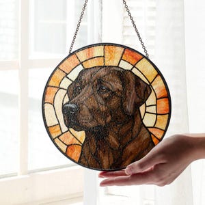 May include: Round stained glass window art featuring a brown Labrador Retriever dog. The dog's portrait is framed by a mosaic of orange and yellow glass. A black metal chain is attached for hanging. The artwork is held by a hand.