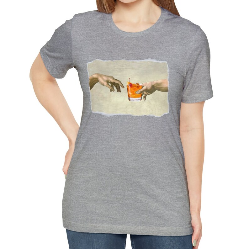 May include: A gray t-shirt with a graphic of two hands reaching towards each other, holding a glass of orange liquid with a cherry on top. The graphic is inspired by the famous Renaissance painting "The Creation of Adam".