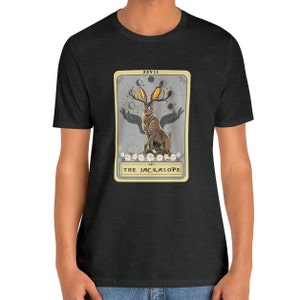 May include: A dark heather grey t-shirt with a graphic of a tarot card featuring a Jackalope, a mythical rabbit with antlers. The card is titled "The Jackalope" and has the Roman numeral XXVII at the top.