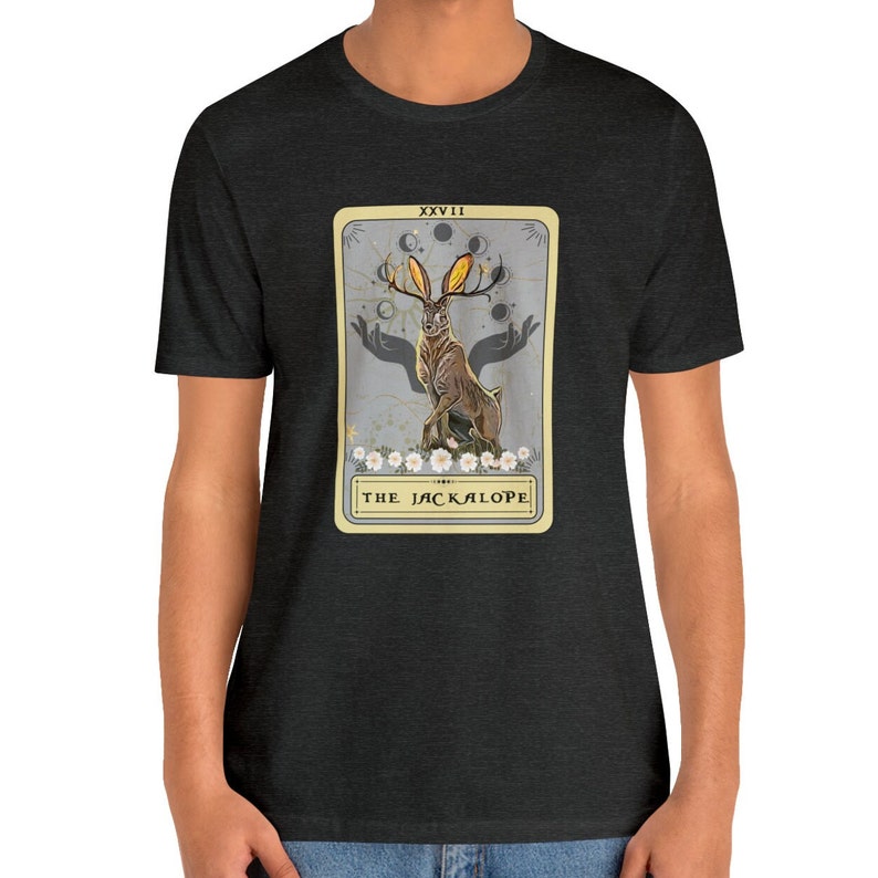 May include: A dark heather grey t-shirt with a graphic of a tarot card featuring a Jackalope, a mythical rabbit with antlers. The card is titled "The Jackalope" and has the Roman numeral XXVII at the top.