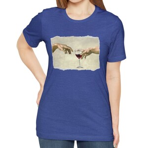 May include: A blue t-shirt with a white distressed rectangle featuring a painting of two hands reaching towards each other, holding a glass of red wine.