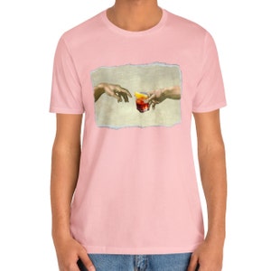 May include: A pink t-shirt with a graphic of two hands reaching towards each other, holding a glass of red liquid with a lemon wedge. The graphic is inspired by the famous Renaissance painting "The Creation of Adam".