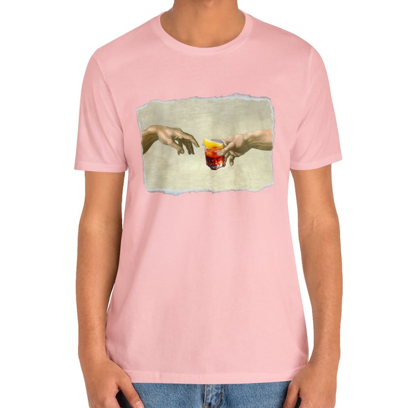 May include: A pink t-shirt with a graphic of two hands reaching towards each other, holding a glass of red liquid with a lemon wedge. The graphic is inspired by the famous Renaissance painting "The Creation of Adam".