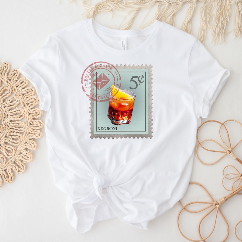 May include: White t-shirt with a vintage-style stamp design featuring a glass of Negroni cocktail and the text "5c" and "NEGRONI".