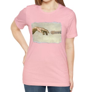 May include: A pink t-shirt with a graphic of a cat's paw and a human hand reaching towards each other, referencing the famous "Creation of Adam" painting.