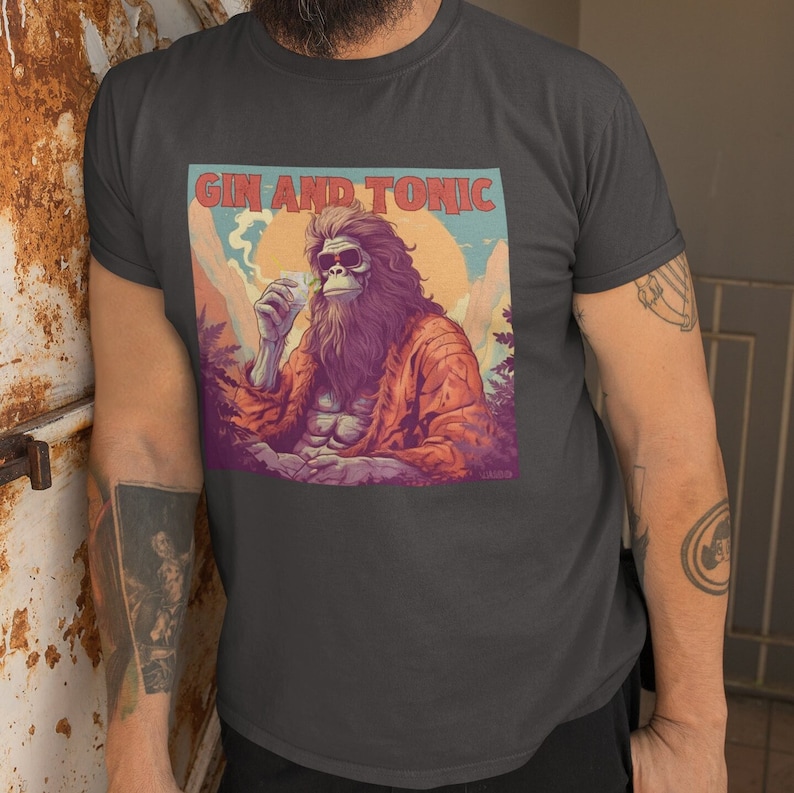 May include: A dark gray t-shirt with a graphic of a hairy, cartoon Bigfoot wearing sunglasses and holding a drink. The text "Gin and Tonic" is above the graphic.