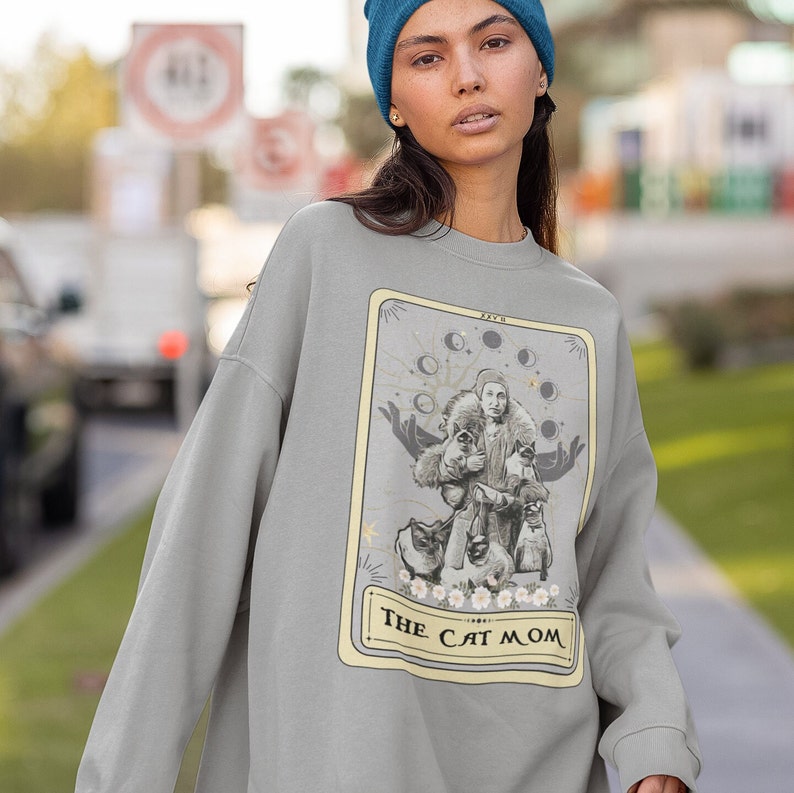 K&ouml;nnte beinhalten: Grauer Pullover mit einem grafischen Druck einer Tarotkarte, auf der eine Frau mit mehreren Katzen und dem Text "THE CAT MOM" abgebildet ist.