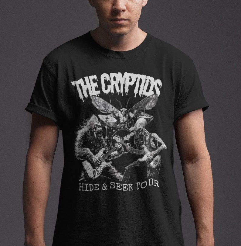 May include: Black t-shirt featuring a white graphic of a band composed of a moth, a Bigfoot, and a skeletal figure playing instruments. The text "THE CRYPTIDS" is above the band, and "HIDE & SEEK TOUR" is below.