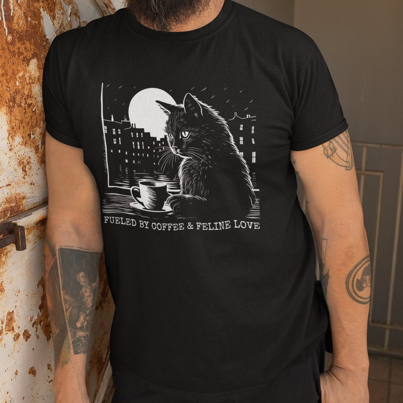 May include: Black t-shirt with a white graphic of a cat sitting by a window with a cup of coffee. The text "Fueled by coffee & feline love" is printed below the image.