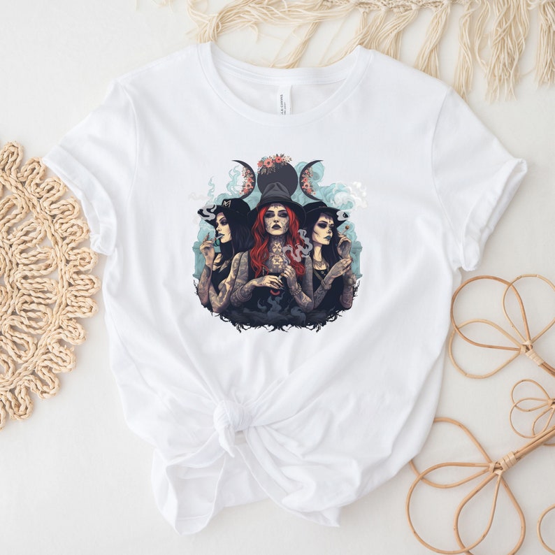 Puede incluir: Camiseta blanca con un gr&aacute;fico de tres mujeres con sombreros de bruja y fumando. Las mujeres est&aacute;n rodeadas de humo y un s&iacute;mbolo de triple luna con flores.