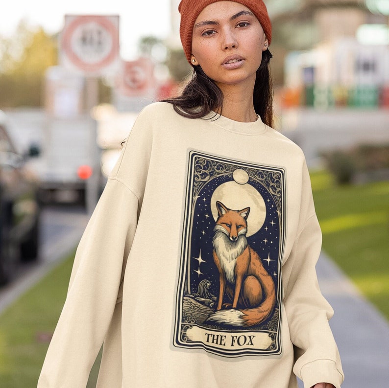 May include: A cream-coloured sweatshirt with a graphic print of a tarot card featuring a fox in front of a full moon. The card reads "THE FOX".