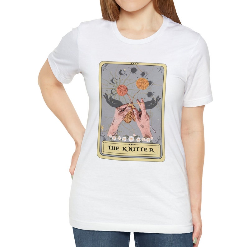 May include: White t-shirt with a graphic of a tarot card featuring a pair of hands knitting with yarn balls and moons in the background. The card reads "The Knitter".