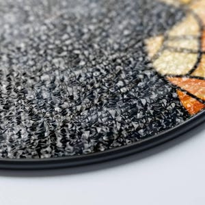 May include: Close-up of a round, textured decorative plate with a black rim. The plate's surface is primarily gray and black with a mosaic-like pattern. Hints of orange and yellow are visible in a section of the plate.