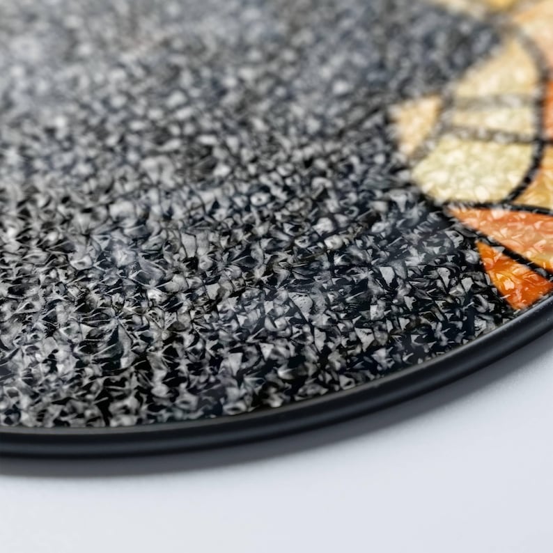 May include: Close-up of a round, textured decorative plate with a black rim. The plate's surface is primarily gray and black with a mosaic-like pattern. Hints of orange and yellow are visible in a section of the plate.
