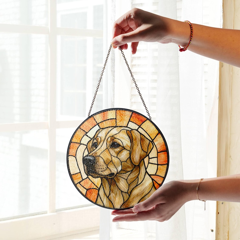May include: Round stained glass window art featuring a Labrador Retriever dog. The dog's portrait is in the center, surrounded by a geometric pattern in shades of orange, yellow, and brown. A black chain is attached for hanging.