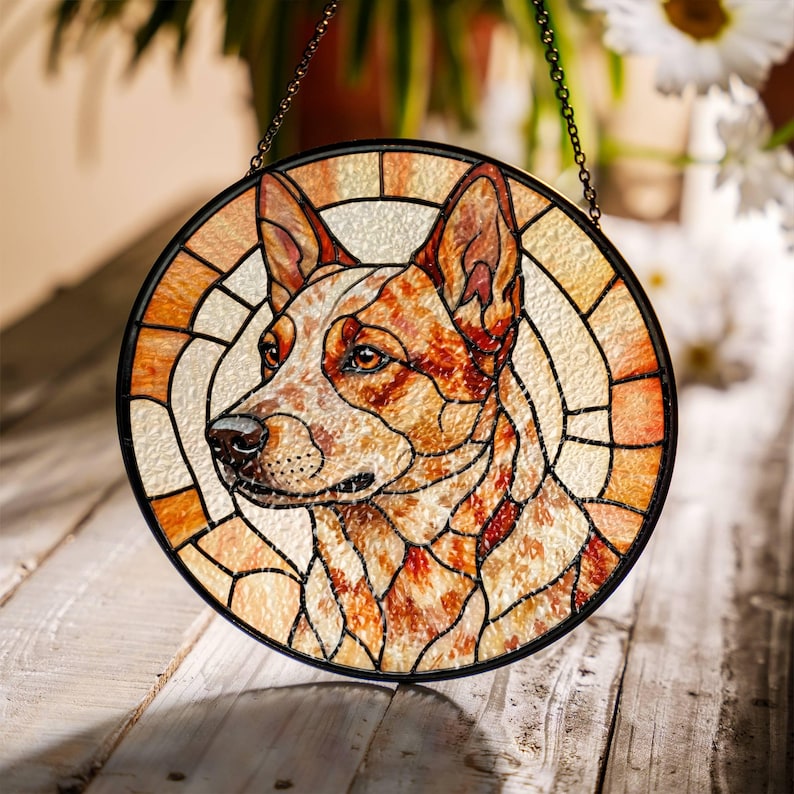 May include: Round stained glass window featuring a dog's portrait. The dog has red and white fur, with a black outline. The glass is segmented with a black border and a chain for hanging. The background is a light beige.