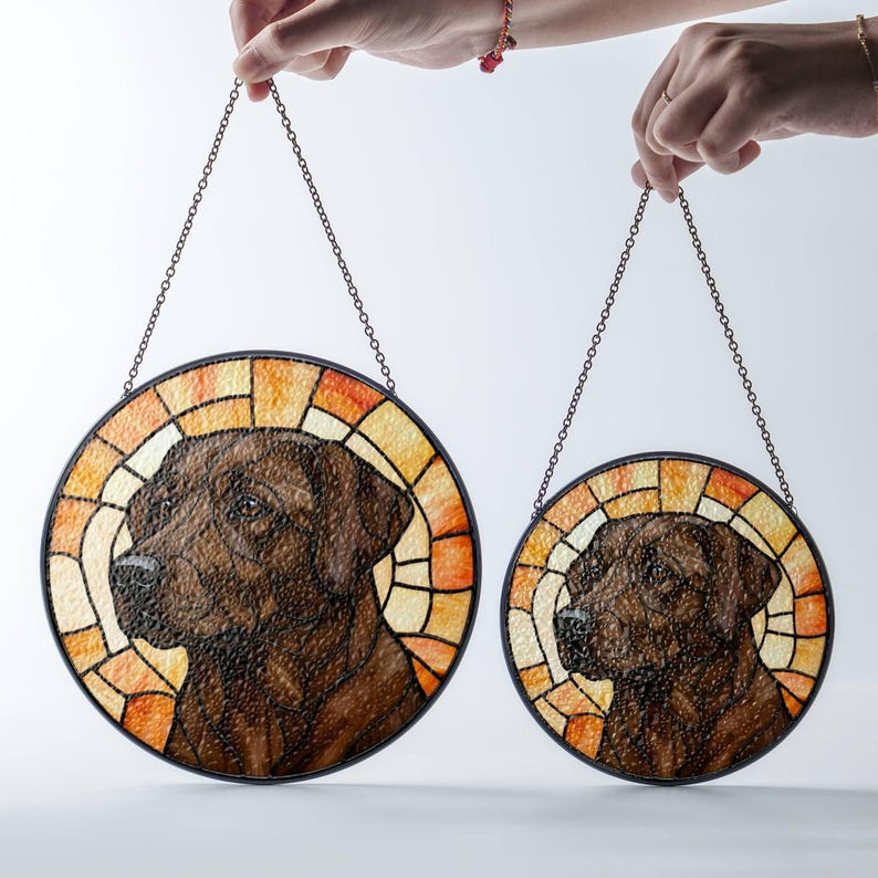 May include: Two round stained glass dog portraits, one large and one small, suspended by chains. The dogs are brown, set against a stained glass background of orange, yellow, and cream. The glass is framed in black.