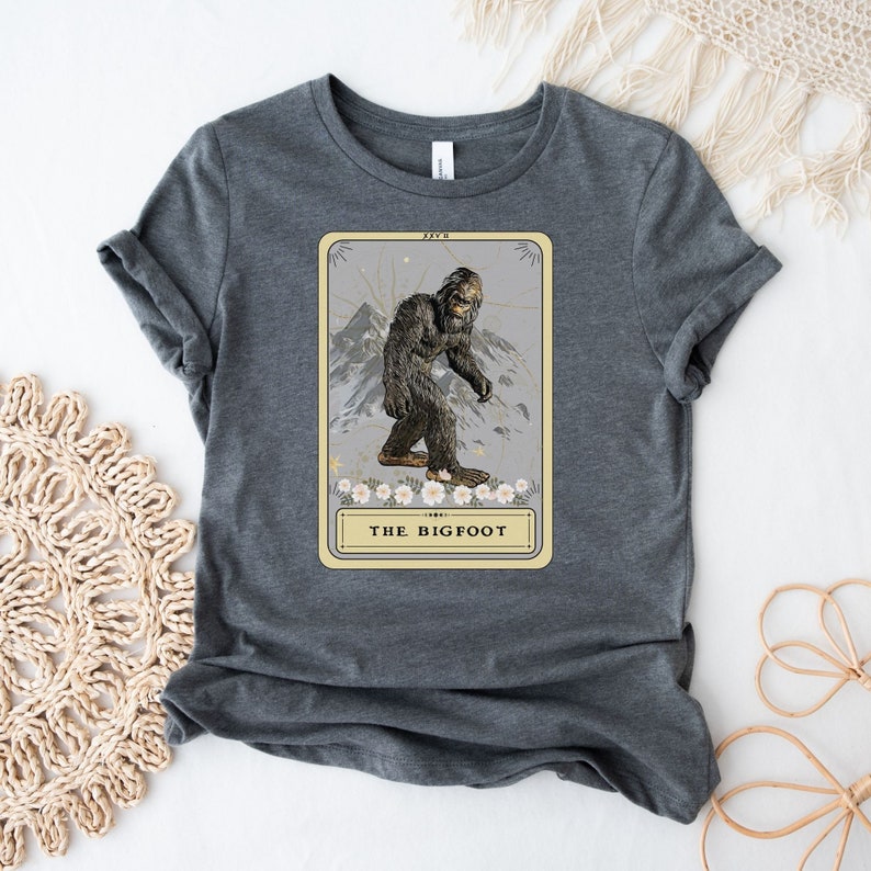Pode incluir: Uma camiseta cinza escura com uma ilustra&ccedil;&atilde;o vintage de Bigfoot em um cart&atilde;o com o texto "THE BIGFOOT".