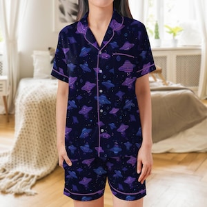Könnte beinhalten: Marineblaues Pyjama-Set mit einem sich wiederholenden Muster aus lila und blauen UFOs. Das kurzärmelige Oberteil und die Shorts sind mit lila Paspeln versehen. Das Oberteil zum Knöpfen hat einen Reverskragen.