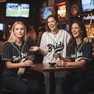 Puede incluir: Tres mujeres en un bar, vistiendo camisetas de béisbol. Una camiseta dice "Bride" en escritura negra, las otras dicen "Team Bride". Las camisetas son a rayas blancas y negras. Se ríen y sostienen bebidas.
