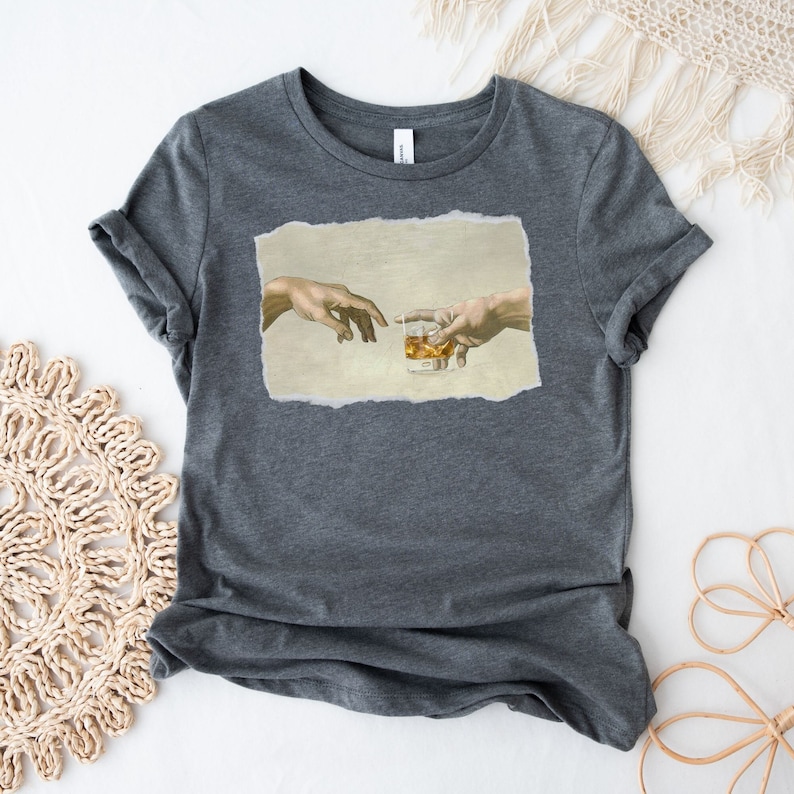 K&ouml;nnte beinhalten: Ein dunkelgraues T-Shirt mit einem Druck des Gem&auml;ldes "Die Erschaffung Adams", aber anstatt dass Gott Adams Finger ber&uuml;hrt, reichen sie beide nach einem Glas Whisky.