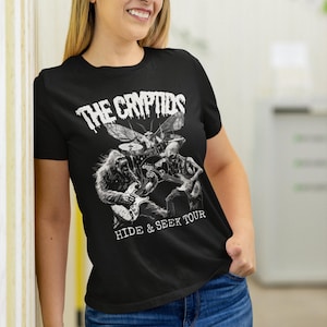 May include: Black t-shirt featuring a band of cryptids, including a Bigfoot-like creature, playing instruments. The band's name, "THE CRYPTIDS," is printed above the band, with "HIDE & SEEK TOUR" below.
