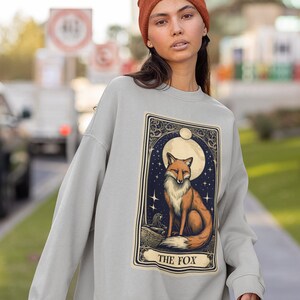 May include: A grey sweatshirt featuring a graphic print of a fox in a moonlit forest. The graphic is in a vintage style and includes the text "THE FOX".