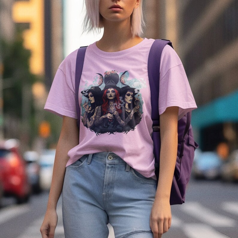 Puede incluir: Una camiseta rosa con un estampado gr&aacute;fico de tres brujas con sombreros y t&uacute;nicas negras, rodeadas de una luna creciente y dise&ntilde;os florales.