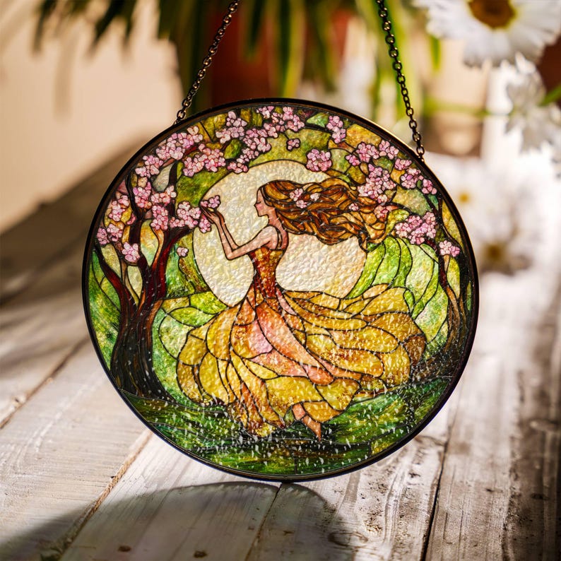 Op de afbeelding: Een hangend glas-in-loodraam toont een vrouw in een vloeiende gele jurk met roze bloemen in haar haar. De vrouw staat in een groen veld met een boom achter haar. Het raam is rond en heeft een metalen frame.