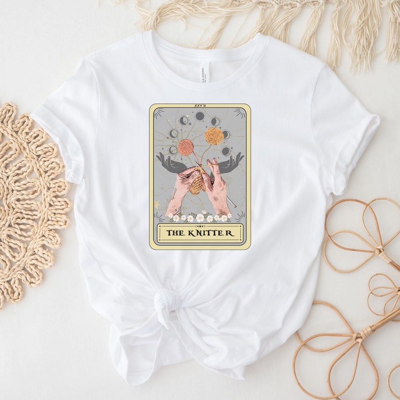 May include: White t-shirt with a graphic of a tarot card featuring a pair of hands knitting with yarn and the text "THE KNITTER".