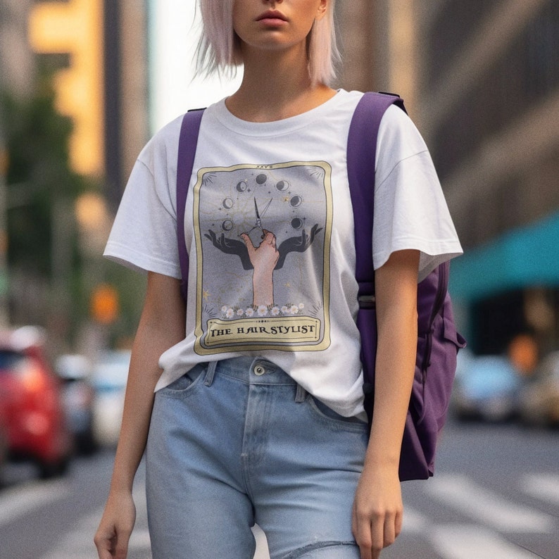 Puede incluir: Camiseta blanca con un gr&aacute;fico de una mano sosteniendo un par de tijeras con una luna creciente y estrellas en el fondo. El texto "THE HAIR STYLIST" est&aacute; impreso debajo del gr&aacute;fico.