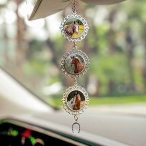 May include: A car charm featuring three circular pendants with images of horses. The pendants are silver-colored with a decorative filigree design. A small horseshoe charm hangs at the bottom. The background is blurred, suggesting an outdoor setting.
