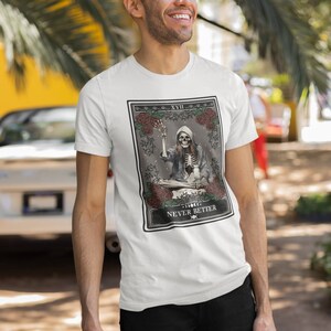 Pu&ograve; includere: T-shirt bianco con una grafica di una carta dei tarocchi che presenta uno scheletro che suona un giradischi. La carta &egrave; etichettata "XVIII" e "NEVER BETTER".