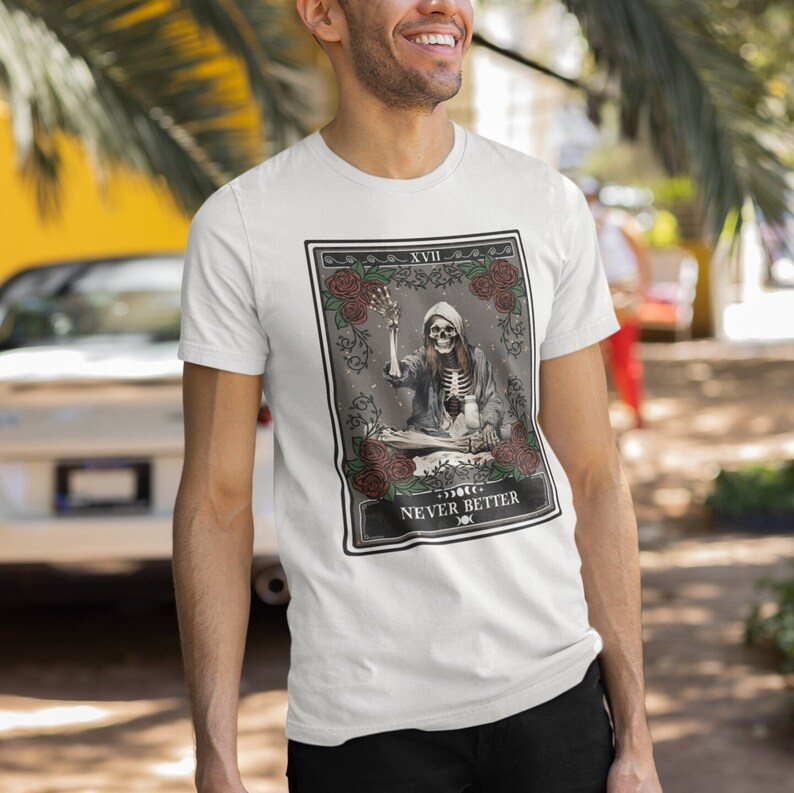 Pu&ograve; includere: T-shirt bianco con una grafica di una carta dei tarocchi che presenta uno scheletro che suona un giradischi. La carta &egrave; etichettata "XVIII" e "NEVER BETTER".