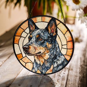 May include: Round stained glass window featuring a dog's portrait. The dog has blue, black, and tan fur, with orange eyes. The window has a black border and a chain for hanging. The background is a mix of orange and cream-colored glass.